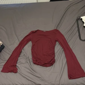 Vinröd långärmad topp Gina Tricot XS - Säljer en vinröd långärmad topp från Gina Tricot i storlek XS. Toppen har figurnära passform, lätt rynkade sidor och vida ärmslut. Perfekt för dig som gillar en enkel men snygg look med lite extra detalj i siluetten.