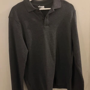 Mörkgrå pikétröja i stickad bomull - Snygg mörkgrå pikétröja med klassisk krage och knäppning framtill. Tröjan är stickad i bomull och har långärmad passform, perfekt för lager på lager. Enkel och stilren design som funkar till många outfits.