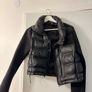 Svart dunväst från Zara - Säljer en svart, quiltad dunväst från Zara med hög krage och dragkedja framtill. Västen har en sportig look och är perfekt att slänga över en hoodie eller tröja. Materialet är syntet och västen har en croppad passform.