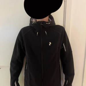 Svart vindjacka Peak Performance S - Svart vindjacka från Peak Performance med huva och dragkedja framtill. Jackan har snygga detaljer med snörning vid halsen och logga på bröstet samt ärmen. Tillverkad i slitstarkt syntetmaterial, perfekt för blåsiga dagar.