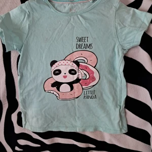 T-shirt med panda och donut (barn) - Söt mintgrön t-shirt med tryck av en panda som chillar i en donut och texten 'Sweet Dreams Little Panda'. T-shirten har korta ärmar och är gjord i mjuk bomull. Perfekt för dig som gillar gulliga motiv och pastellfärger. Jätte fint skick- behöver bara tvättas.