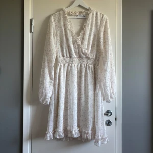 Vit blommig omlottklänning från Vila - Supersöt vit kortklänning från Vila med ljusrosa blommönster, omlott och v-ringning med volangdetaljer. Klänningen har långa ärmar, smock i midjan och volangkant nertill. Perfekt för sommarens alla tillfällen! Endast använd 2 gånger 