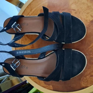 Svarta sandaler med klack och spänne - Snygga svarta sandaler med kilklack och remmar i mockaimitation. Spänne i silverfärg vid ankeln och flätad sula i repstil ger en somrig vibe. Perfekta nu till sommaren. Bra skick- använde de endast 2 gånger, passar inte mig längre.