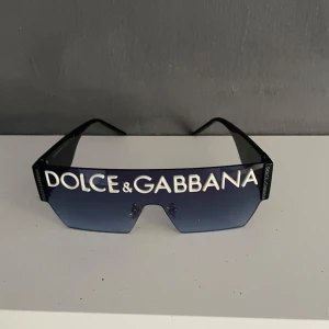 Dolce & Gabbana svarta solglasögon - Snygga svarta solglasögon från Dolce & Gabbana med stor logga framtill och DG på sidorna. Sköldformade glas med mörkblå toning och breda skalmar.  Passar både till kille eller tjej 