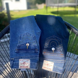 True Religion blå bootcut jeans - Säljer ett par ikoniska blå jeans från True Religion med snygga kontrastsömmar och stora fickor med lock och knapp bak. Jeansen har låg midja och bootcut-ben, klassisk logga på bakfickan och coola detaljer. Använda med är i bra skick i storlek 24.