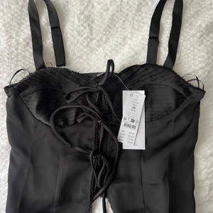 Svart satin topp med knytdetaljer framtill - Supersnygg svart topp i satin från Gina Tricot. Toppen har breda axelband såsom går att justera,  snörning framtill och veckade detaljer över bysten. 