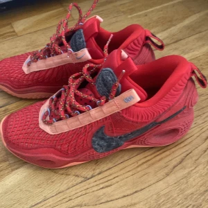 Basketskor - Unika röda Nike sneakers med svart swoosh och coola detaljer. Skorna har mesh- och mockaliknande material, färgglada snören och rosa paneler längs sidorna. Perfekta för dig som vill sticka ut med en sportig och trendig look. Nike cosmic unity 2