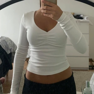 Vit långärmad topp med v-ringning - Säljer en vit långärmad topp med v-ringning och snygg rynkdetalj framtill. Modellen är figurnära och har en l om, stilren look som passar perfekt till jeans eller kjol. Mjuk och stretchig i materialet för en bekväm känsla. 