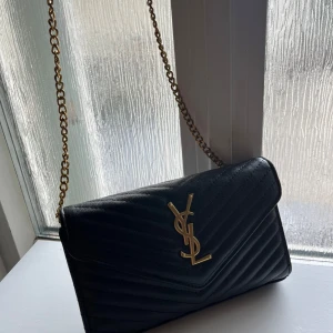 Svart quiltad YSL axelväska i skinn - Snygg svart axelväska från Saint Laurent med guldiga detaljer och kedjerem. Väskan är quiltad i ett chevronmönster och har YSL-logga framtill. Tillverkad i skinn och har flera fack inuti, inklusive ett med dragkedja. Perfekt för dig som gillar lyxiga accessoarer. AA copy