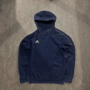 Mörkblå Adidas hoodie med huva - Mörkblå hoodie från Adidas med broderad logga på bröstet. Tröjan har huva, långa ärmar och en stor magficka, perfekt för streatwear men också andra stilar! 