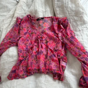 Blommig volangblus  - Superfin blommig blus i rosa, lila och röda toner. Tunn och luftig med volangdetaljer, v-ringning och långa ärmar. Perfekt för dig som gillar färg och romantiska vibes.  i storlek S.
