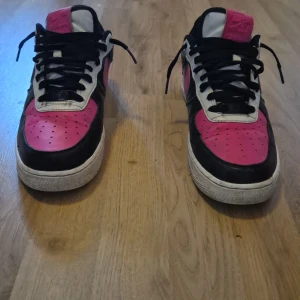 Nike Air Force 1 svart/rosa custom - Unika Nike Air Force 1 sneakers i svart och rosa med vita detaljer. Klassisk låg modell med svart skinn, rosa swoosh och tåparti samt vita inslag runt snörningen och hälen. Svarta skosnören och vit sula med rosa AIR-tryck. Perfekta för dig som vill sticka ut.