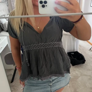 Mörkgrå blus med smock och v-ringning - Supersöt mörkgrå blus med v-ringning och knytband. Smockad midja med broderade detaljer och lätt volang nertill. Korta ärmar och luftig passform, perfekt till jeans eller kjol. Ger en avslappnad men ändå chic vibe.