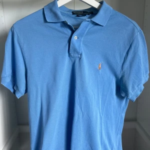 Blå pikétröja från Ralph Lauren - Klassisk ljusblå pikétröja från Ralph Lauren med broderad logga på bröstet. Tröjan har krage, korta ärmar och knäppning med två knappar framtill. Tillverkad i bomull för en skön och luftig känsla. Perfekt för en clean och sportig stil.