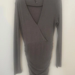 Glittrig grå omlottklänning BikBok XS - Supersnygg grå kortklänning från BikBok i omlottmodell med glittrigt tyg. Klänningen har lång ärm, v-ringning och snygg drapering framtill. Perfekt för dig som vill sticka ut med en stilren men ändå festlig look!