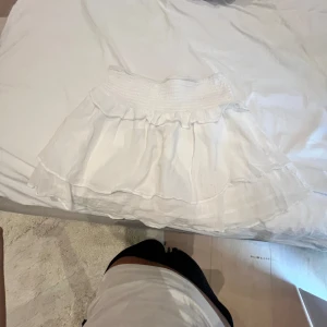 Vit volangkjol från Gina Tricot - Supersöt vit kortkjol från Gina Tricot med bred smock i midjan och dubbla volanger. Kjolen är luftig och lätt, perfekt för varma dagar. Tillverkad i mjukt bomullstyg som känns skönt mot huden. Enkel att matcha med både toppar och sneakers.