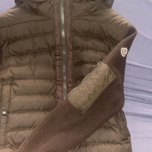 Brun Moncler dunjacka med stickade ärmar - Säljer en brun Moncler dunjacka med stickade ärmar och detaljer i samma färg. Jackan har dragkedja framtill, quiltad kropp och Moncler-logga på ärmen. Perfekt för dig som vill ha en snygg och varm jacka med unik design och premiumkänsla.