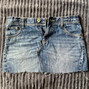 Blå jeanskjol med rå kant - Säljer en blå jeanskjol i klassisk denim med slitna detaljer och rå, fransig kant nertill. Kjolen har fem fickor, bälteshällor och dragkedja med knapp framtill. Perfekt för dig som gillar en avslappnad och cool stil.