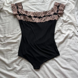 Svart body med spets från Intimissimi - Supersnygg svart body från Intimissimi med bred, ljusrosa spets upptill och offshoulder-modell. Kroppen är slät och figurnära, medan spetsen ger en romantisk vibe. Perfekt att styla med jeans eller kjol för en trendig look.
