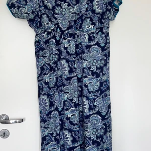 Mönstrad långklänning i blått - Snygg långklänning med paisleyinspirerat mönster i olika blå toner och inslag av vitt och turkost. Klänningen har korta ärmar och resår i midjan för en bekväm passform. Perfekt för dig som gillar bohemisk stil och vill sticka ut med ett coolt print.