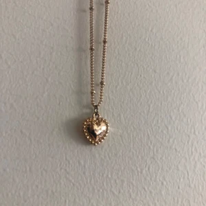 Guldigt halsband med hjärthänge - Supersött halsband i guldton med ett hjärtformat hänge. Hänget har en pärlad kant som ger extra detalj och kedjan är tunn och elegant. Perfekt accessoar för att lyfta vilken outfit som helst.