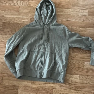 Grå hoodie med dragkedja från H&M - Säljer en basic grå hoodie från H&M med dragkedja framtill och huva med snörning. Tröjan har två stora fickor på magen och är tillverkad i mjuk bomull. Perfekt för chill dagar eller när du vill ha en enkel och stilren look.