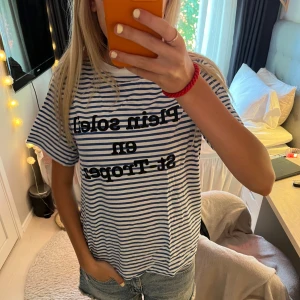 Randig t-shirt med St-Tropez-tryck - Säljer en vit och blå randig t-shirt med svart texttryck 'Plein soleil en St-Tropez' på bröstet ifrån H&M. T-shirten har korta ärmar, rund hals och en avslappnad passform. 