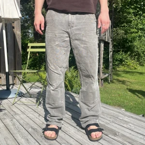 Grå cargopants från Carhartt - Säljer ett par grå cargopants från Carhartt med flera praktiska fickor och coolt, tvättat utseende. Byxorna har raka ben och en avslappnad passform. Perfekta för dig som gillar streetwear och funktionella detaljer. Klassisk Carhartt-logga på bakfickan. OBS, ena fickan har ett stort hål i sig och tyget börjar sakta spricka i skrevet
