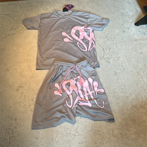Syna world - Säljer ett par Syna world  med ett stort rosa graffiti-inspirerat tryck på framsidan. T-shirten har korta ärmar och en avslappnad passform. Materialet känns mjukt och sportigt, perfekt för dig som gillar streetwear och vill sticka ut med coola detaljer.