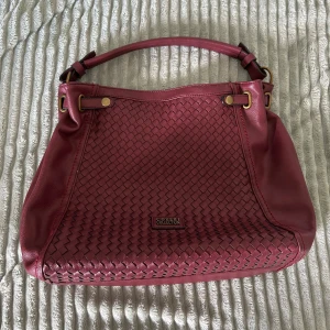 Vinröd axelväska från Coveri Collection - Snygg vinröd axelväska i skinn från Coveri Collection med flätad framsida och guldfärgade metalldetaljer. Väskan har både handtag och avtagbar axelrem, flera fack med dragkedja och ett rymligt innerfoder med fickor.