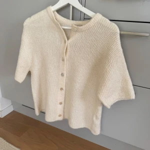 Krämvit stickad kofta från Gina Tricot premium collection - Supermjuk och ribbstickad kofta i krämvit färg från Gina Tricot. Kortärmad modell med rund halsringning och knäppning framtill. Från deras premium collection.