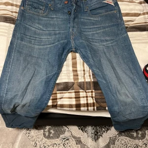 Replay blå jeans, storlek 33 - Säljer ett par klassiska blå jeans från Replay i storlek 33. Jeansen har snygga slitningar, fem fickor och coola detaljer på bakfickorna. De är tillverkade i jeansmaterial och har en rak passform som funkar till det mesta.