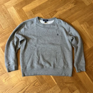 Grå sweatshirt från Polo Ralph Lauren - Klassisk grå sweatshirt från Polo Ralph Lauren med broderad marinblå logga på bröstet. Tröjan har rund halsringning, långa ärmar och är tillverkad i mjuk bomullsblandning. Perfekt för en chill och stilren look.