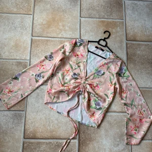 Blommig omlottopp från BikBok - Superfin blommig omlottopp från BikBok i ljusrosa med gröna, lila och vita detaljer. Toppen har lång ärm, v-ringning och snörning framtill som ger en snygg rynkad effekt. Tunt, lätt material som känns luftigt och perfekt för vår och sommar. Vet inte om de är nån ”missfärgning” på sista bilden men tror den var sån från butik! 