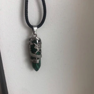 Halsband med grön sten och drake - Unikt halsband med en spetsig grön sten omsluten av en detaljerad silvrig drake. Hängs i ett svart snöre. Perfekt för dig som gillar fantasy eller vill ha något annorlunda.