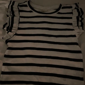 Randig topp från Uniqlo Studio - Säljer en vit och svart randig topp från Uniqlo Studio. Toppen har volangdetaljer på axlarna och är ärmlös, vilket ger en cool och trendig look.Materialet känns mjukt och bekvämt.Storlek 158/164 men passar mig som har S! väldigt bra skick!💘