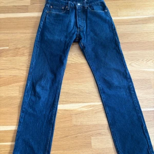 Levi's 501 blå jeans straight fit - Klassiska Levi's 501 jeans i mörkblå denim med raka ben och normal passform. Byxorna har fem fickor, knappgylf och den ikoniska läderpatchen bak i midjan. Perfekta för dig som gillar tidlös stil och schysst kvalitet.
