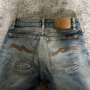Slim nudie Jeans - Snygga blå skinny jeans från Nudie Jeans med klassisk orange söm på bakfickorna och läderpatch i midjan. Jeansen har en tight passform, normal midja och slitna detaljer för en cool look. Perfekta för dig som gillar en smal siluett.