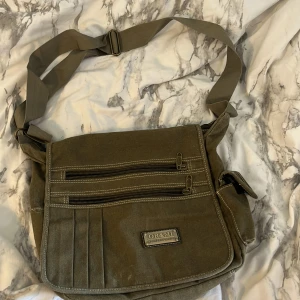 Olivgrön axelväska från OR&MI - Olivgrön axelväska från OR&MI i slitstark bomull med justerbar rem. Väskan har flera dragkedjefack framtill och på insidan, samt en praktisk sidoficka. Perfekt för att bära laptop, böcker eller vardagsprylar. Snygga kontrastsömmar och metallmärke framtill. Köpt utomlands tror Malta, jätte fin men kommer tyvärr inte till användning längre ❤️