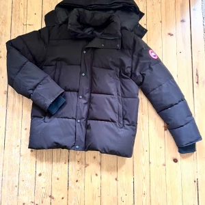 Canada goose jacka  - Säljer en svart dunjacka från Canada Goose med klassisk patch på ärmen. Jackan har hög krage, avtagbar huva och stängs med både dragkedja och knappar. Fodrad för extra värme och har långa ärmar med muddar. Perfekt för kalla dagar. Hängaren har gått sönder tyvärr. Storlek XL