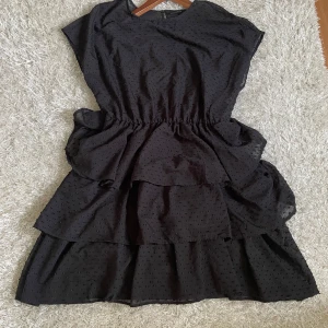 Svart prickig volangklänning Gina Tricot - Supersöt svart klänning från Gina Tricot med små prickar i tyget och volangdetaljer i kjolen. Modellen är kort och har en lös passform med markerad midja. Perfekt för dig som gillar en trendig stil.