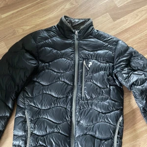 Svart dunjacka Peak Performance  - Svart dunjacka från Peak Performance med vågformat quiltat mönster. Dragkedja framtill. Perfekt för kyliga dagar och snygg att bära till det mesta.