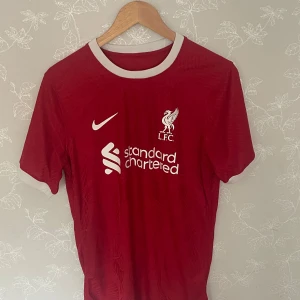 Liverpool FC hemmatröja 2023/2024 - Säljer en röd Liverpool FC hemmatröja från Nike med vita detaljer och klubbmärke på bröstet. Tröjan har korta ärmar och sponsorlogga framtill. Perfekt för dig som älskar fotboll och vill visa ditt stöd för LFC!