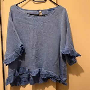 Blus strl S  - Blå blus med volanger.  Det står inte storlek men personen på bilden har vanligtvis storlek XS så skulle säga att blusen är en S om man inte vill att den ska sitta löst.  Material: linne och bomull 
