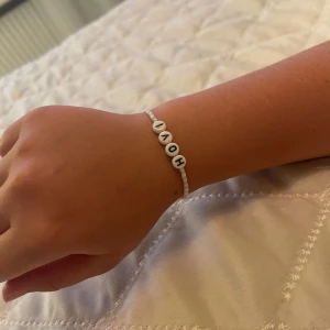 Pärlarmband med texten HOV1 - Ett sött armband med små vita och ljusblå pärlor samt fyra större vita pärlor med svarta bokstäver som bildar ordet HOV1. Perfekt accessoar för dig som gillar personliga smycken och vill sticka ut lite extra.