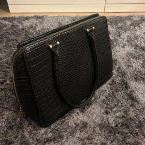 Svart väska i croco-imitation - Snygg svart väska i croco-imitation med guldiga detaljer och dragkedjor. Väskan har flera fack och ett coolt mönstrat innerfoder. Perfekt för att bära laptop och allt du behöver till plugget eller jobbet.