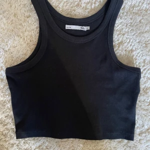 Svart croppad linnetopp  - En svart croppad linnetopp från lager 157  med sportig skärning och breda axelband. Toppen är ribbad och har en tight passform som sitter snyggt mot kroppen. Perfekt att styla med jeans eller kjol för en clean look.