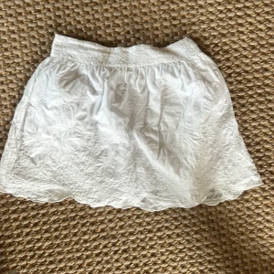 Vit broderad kortkjol med volangkant - Superfin vit kortkjol med broderade detaljer och vågig volangkant nertill. Kjolen har en bred, smockad midja som ger en bekväm passform. Perfekt för varma dagar och ger en fräsch och somrig vibe.