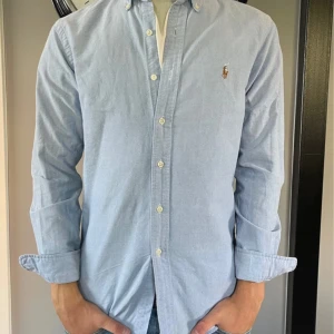 Ljusblå skjorta från Ralph Lauren - Snygg ljusblå skjorta från Ralph Lauren i slim fit-modell. Skjortan har klassisk krage med knappar, lång ärm och broderad logga på bröstet. Tillverkad i mjuk bomull och passar perfekt till jeans eller chinos. Pris är inte hugget i sten.🪨 🗡️