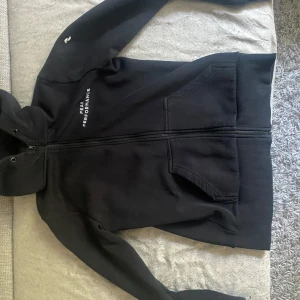 Svart hoodiejacka från Peak Performance - Svart hoodiejacka från Peak Performance med dragkedja framtill och två fickor. Jackan har huva, ribbade muddar och logga på bröstet. Knappt använd passar storlek M